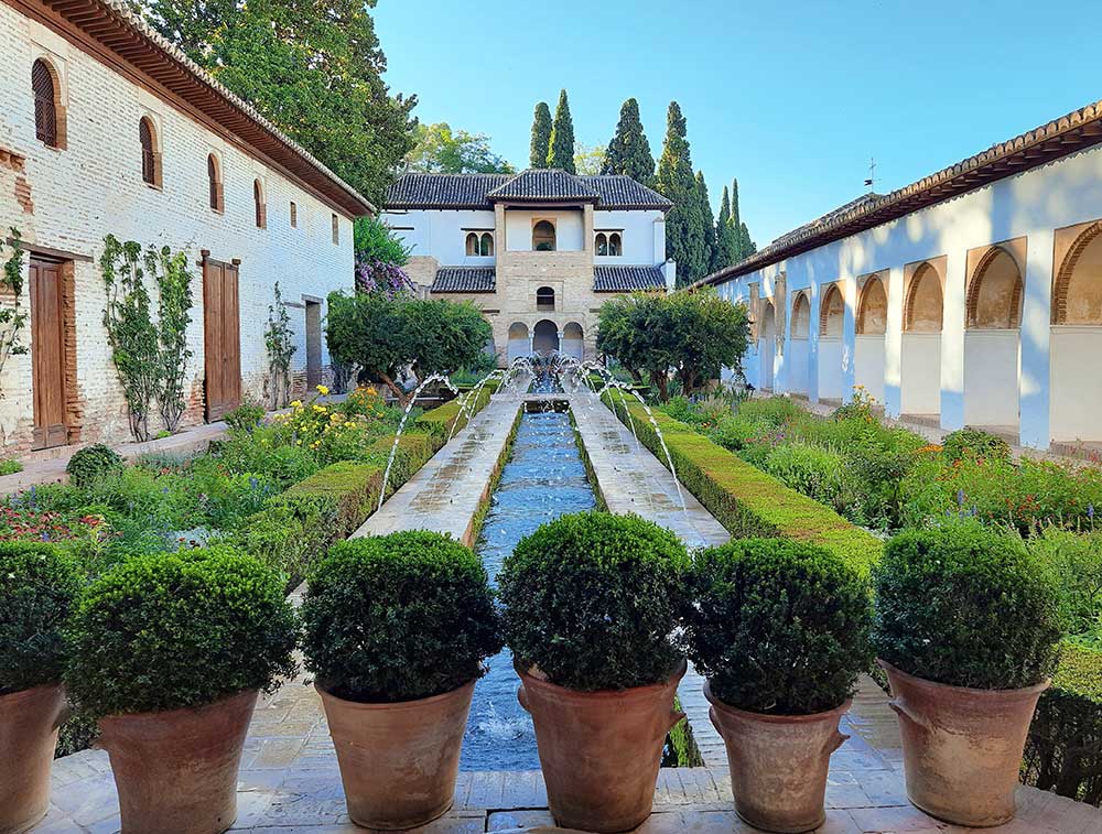 The Water Garden Courtyard, a must see on your Granada, Spain Itinerary featuring a long rectangle pond with fountain surrounded by well-manicured garden of plants and trees