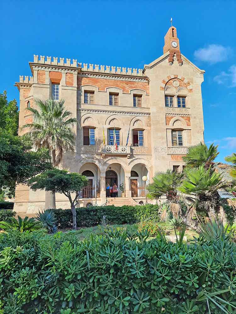 A large neo-gothic and neopolitan style house in a pinkish orange colour with manicured gardens in front. 