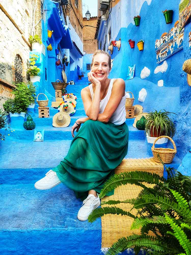 A woman (me) wearing a white sleeveless top and a long green skirt is sitting on the blue street in Chefchaouen - this maxi skirt came to just above my ankles and was the perfect length to wear in Morocco.