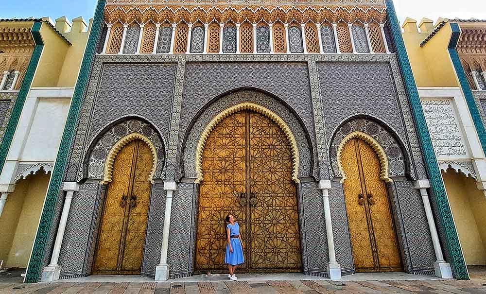 Image of Fez Palace in Morocco with three ornate doors and a woman standing in front of the middle one. Fes is up there as one the most beautiful city in Morocco = read this article and decide for yourself