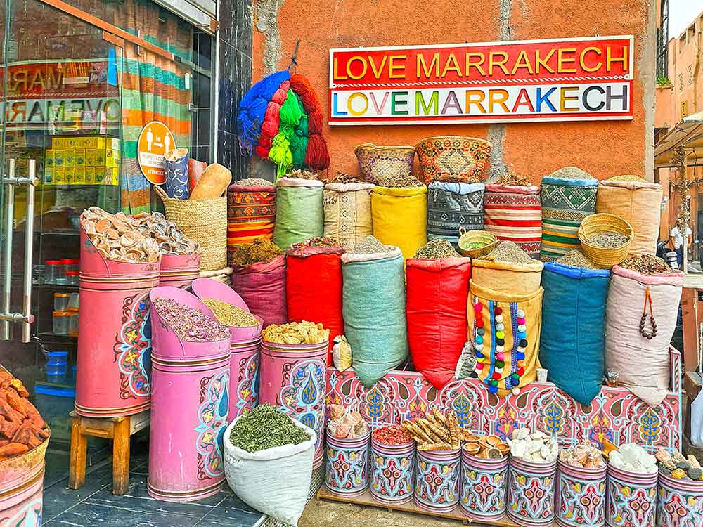 A shop stall in Marrakech. There are piles of spices in colourful bags stacked up aggainst a wall. On the wall it says 'love marrakech' self-guided 10 days Morocco itinerary from Casablanca to Marrakech