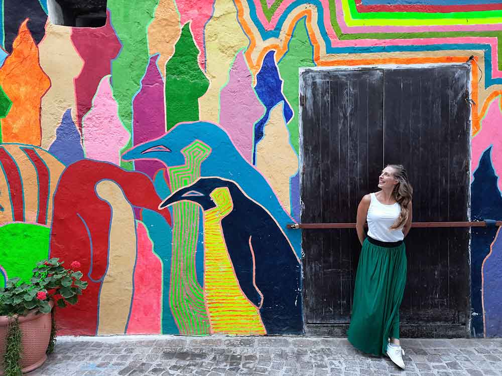 A woman (me) wearing a white sleeveless top and a long maxi skirt, along with comfortable white shoes, stands near one of the street art displays in Essaouira
