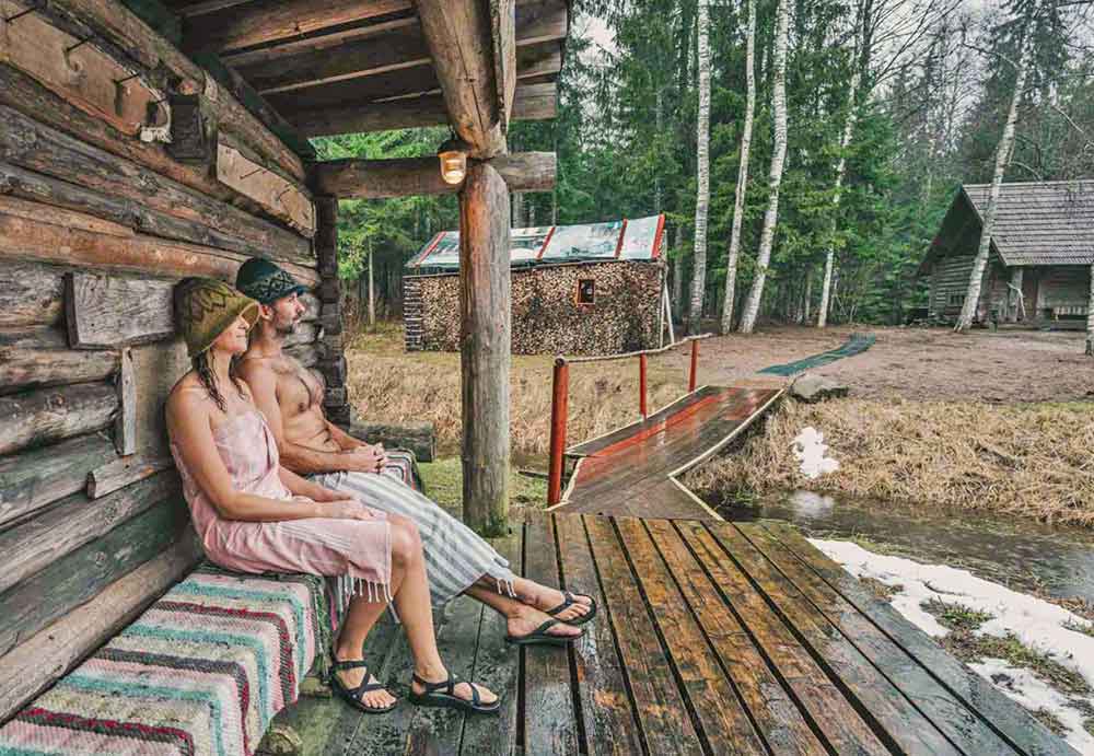 A couple sitting on the side of the house took a Mooska Farm Smoke Sauna in a charming log cabin surrounded by lush forests, meadows, and pristine nature.