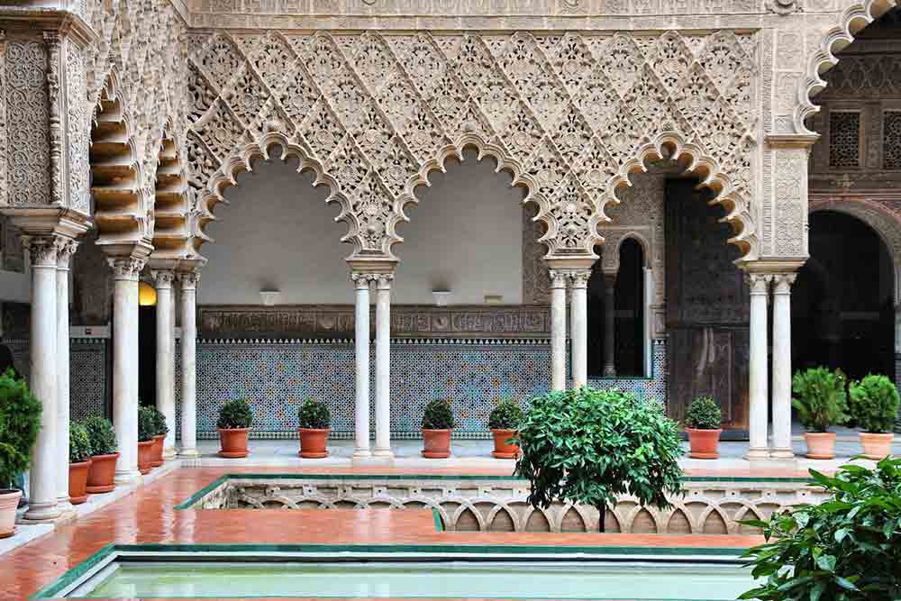 The Real Alcazar in Seville features arches with intricate details, mosaic walls, and rows of plants.