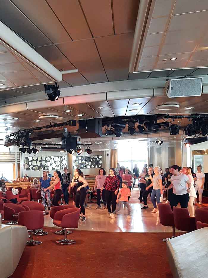 A group of people learning to dance. There is a circular dance floor surrounded by red chairs and sound and audio equipment on the ceiling.