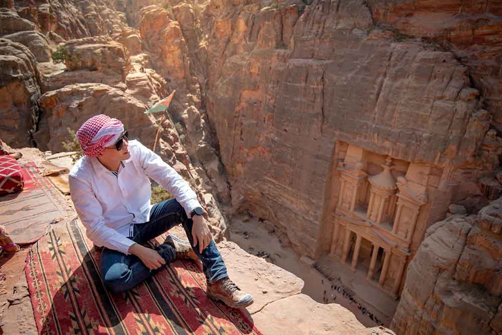 Man with a white long-sleeve and pants sitting on carpet viewpoint in Petra Ancient City looking at the Treasury