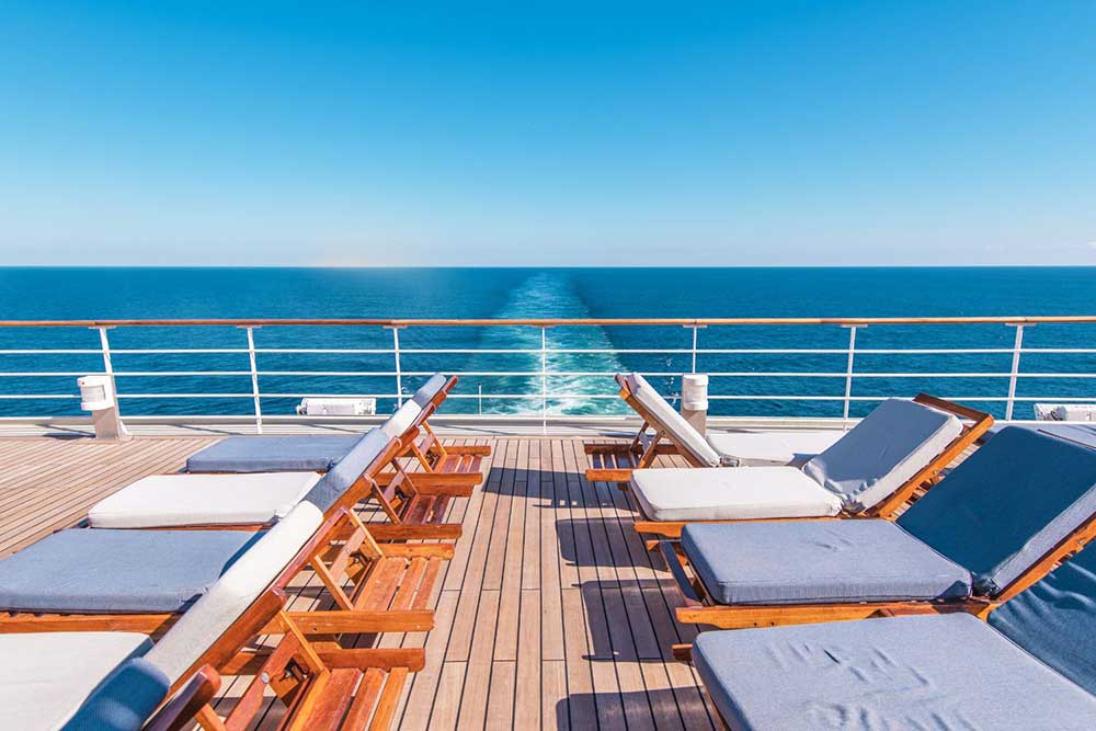 Sun beds with blue and white cushions on the deck of a cruis ship. There is a raling and the waves that the ship has left. The sea is deep blue the sky is lighter blue. 