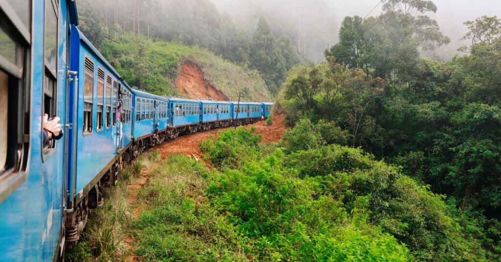 Image of a blue train in Sri Lanka riding through the countryside with hills and trees. Taking a ride on the Kandy to Ella train is one of the top Sri lanka bucket list things to do in Sri lanka.