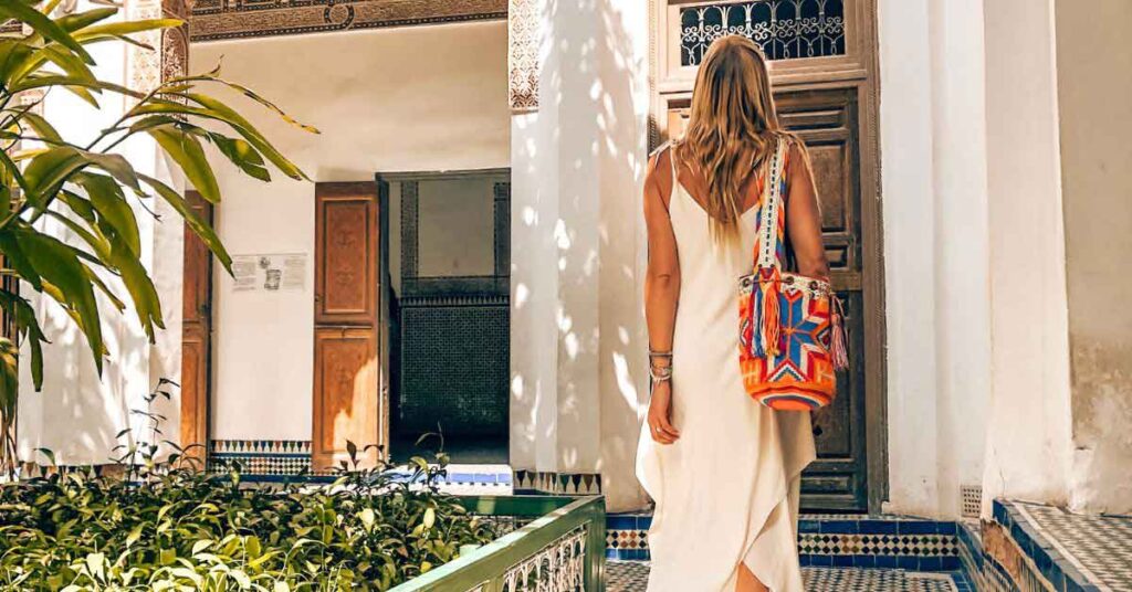 A woman with blonde hair looking away from the camera wearing a long white linen dress in Marrakech, shes carrying a colourful shoulder bag and admiring the buildings. This is the featured image for an article about what to wear in Marrakech.