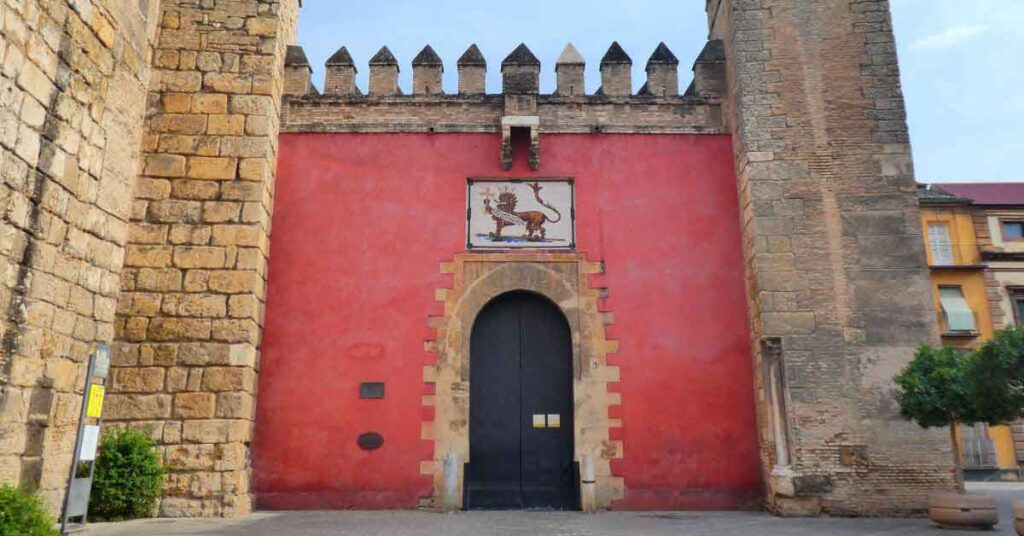 Puerta del León (Real Alcázar de Sevilla), the main entrance gate with a striking red wall and a tile of a lion above the arched doorway, leading to the palaces and gardens inside the Real Alcázar of Seville.