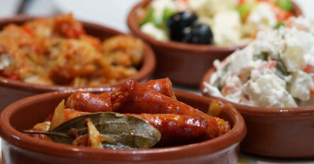 Close-up of assorted Spanish tapas in terracotta dishes, including chorizo with bay leaves, creamy potato salad, and vegetable-based starters. This image captures the vibrant flavors featured in a Seville tapas tour review, showcasing the variety experienced on a walking food tour with Devour Tours.