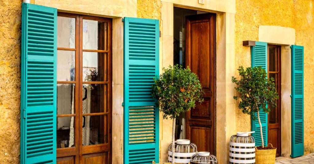 A rustic Mediterranean-style house with bright turquoise shutters, wooden doors, and potted plants in the sun. The warm stone walls and soft lighting create a relaxed atmosphere, evoking the feeling of winter sun in Europe.